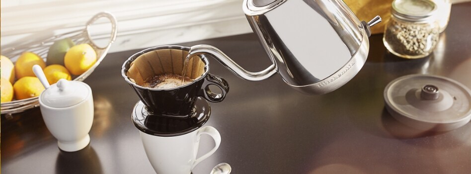 Someone using a KitchenAid Electric Gooseneck Kettle to pour hot water into a pour over cone on a mug. Also on the counter is a basket filled with lemons and limes, a sugar bowl, a potted plant, two containers and the base of the kettle.